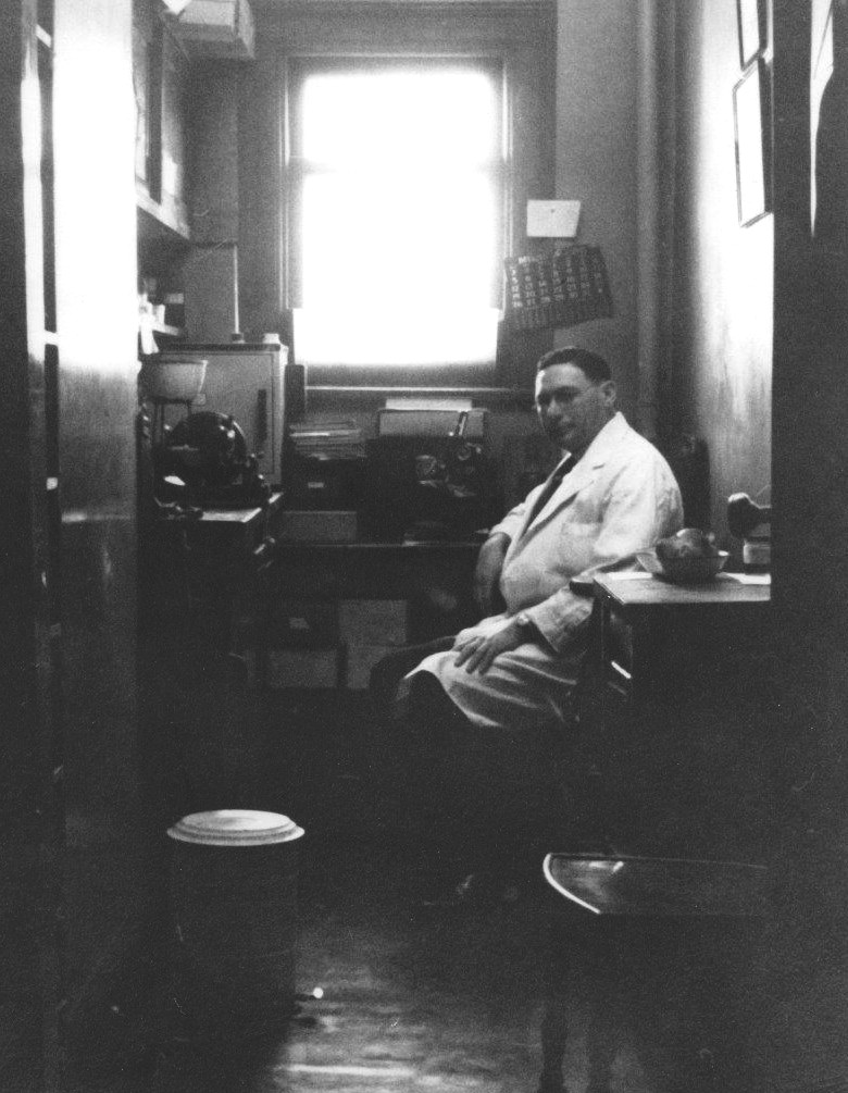 This image shows Dr. Harold Pritzker seated in a small room with a window. A microscope and other equipment sits on tables surrounding him. 