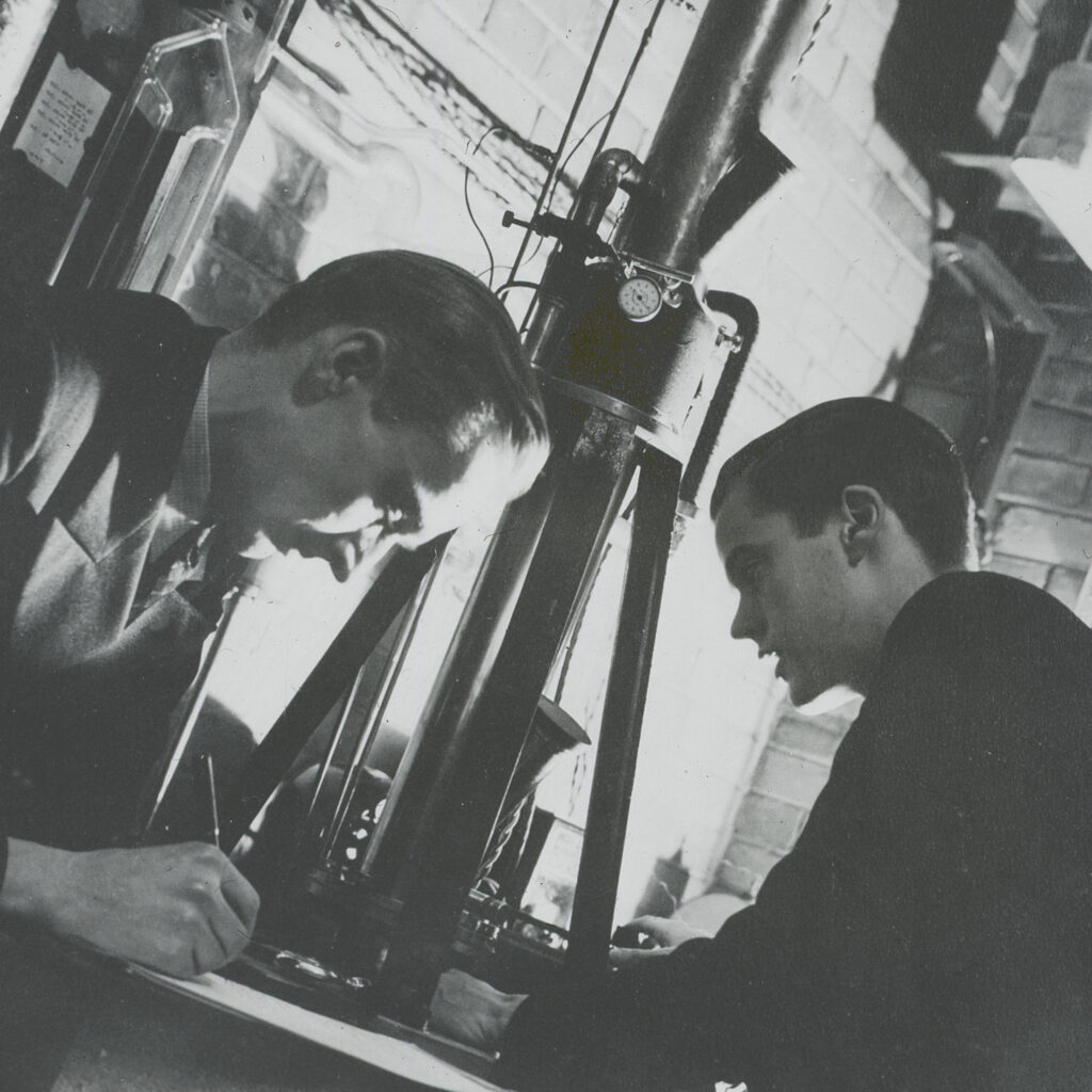 A cropped section of a black-and- white photograph, likely from 1938, showing Albert F. Prebus (1913-1997) and James Hillier (1915-2007) in front of the recently-built electron microscope.