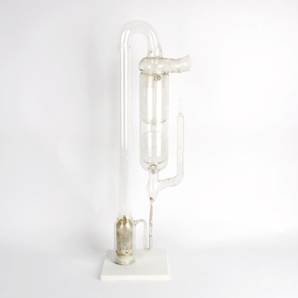 A complex glass instrument set on a rectangular white stand. A fluid reservoir at the base of the artifact joins a long tube at its top. A curve in this tube forms the uppermost part of the instrument. This curve broadens into a complex element consisting of the pump inlet, the mercury vapour jets, and a water jacket for condensing the mercury. Below this is the pump exhaust and a narrow tube for returning the condensed mercury to the fluid reservoir. 